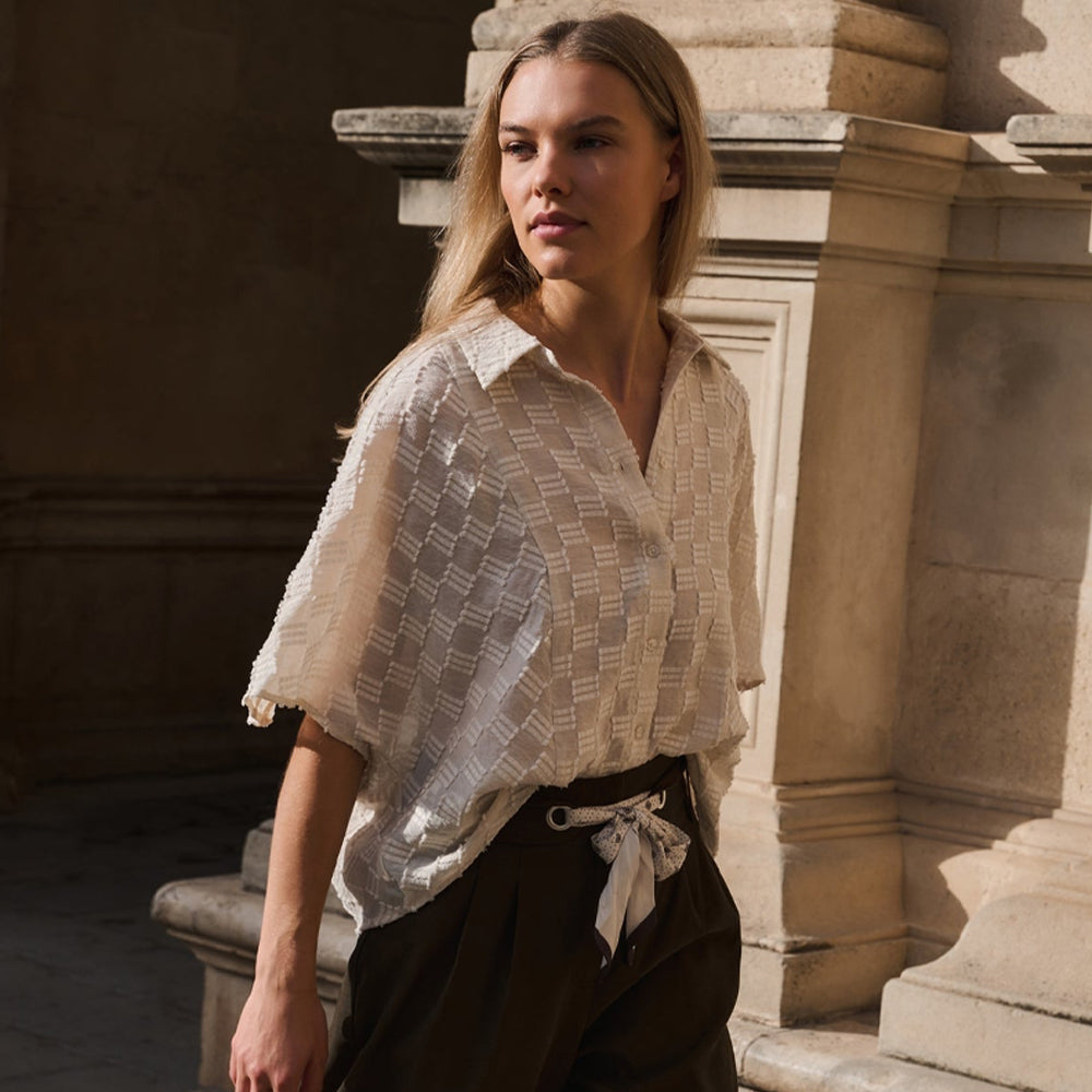 A woman wearing a cream-colored textured short-sleeve button-up blouse and dark brown trousers, standing in front of a stone architectural column.