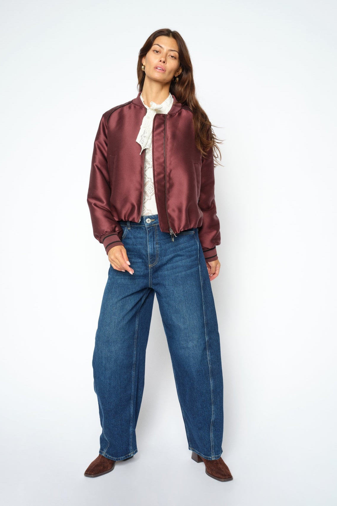 A woman modeling a cropped satin bomber jacket in burgundy, paired with wide-leg blue jeans, a white ruffled blouse, and brown shoes, against a plain white studio background.