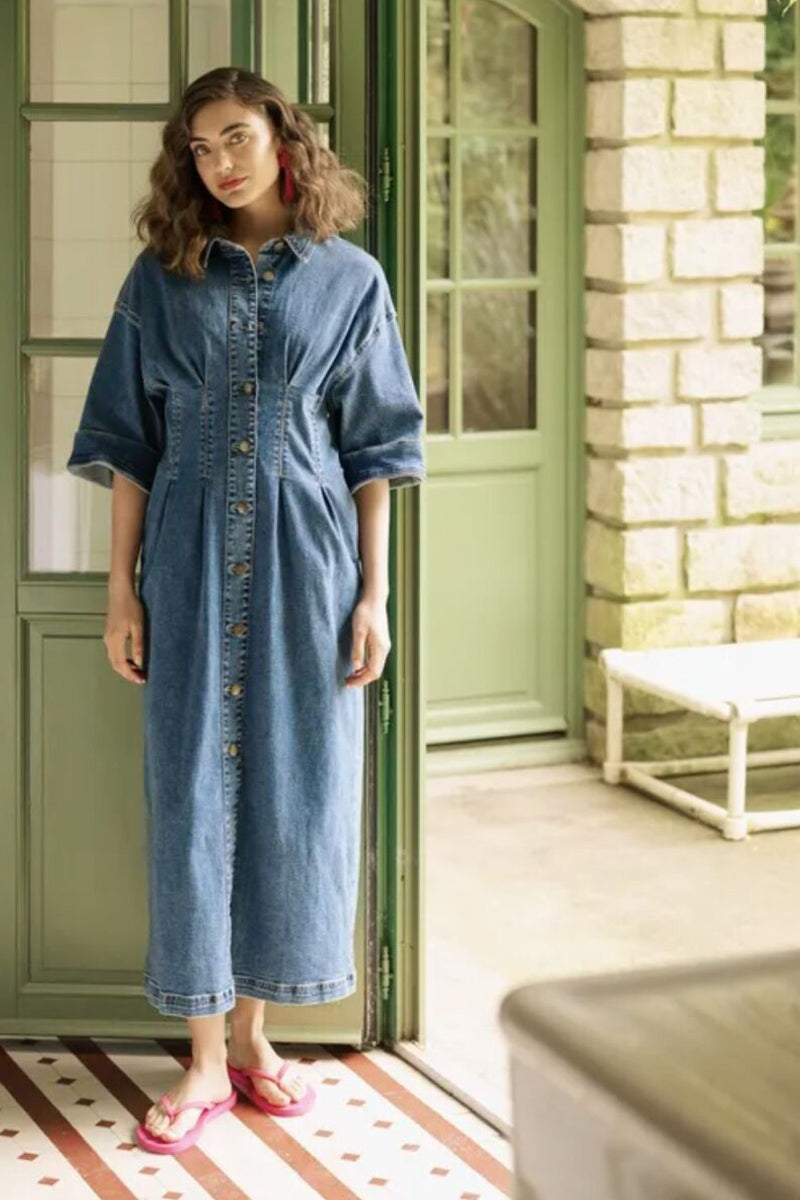 Woman wearing a blue denim dress standing in a room with green doors and a stone wall.