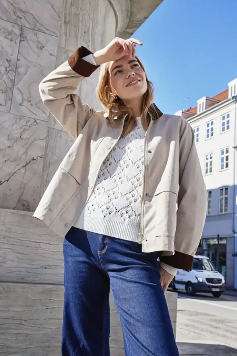 Woman in beige jacket and white sweater standing outdoors on a sunny day.